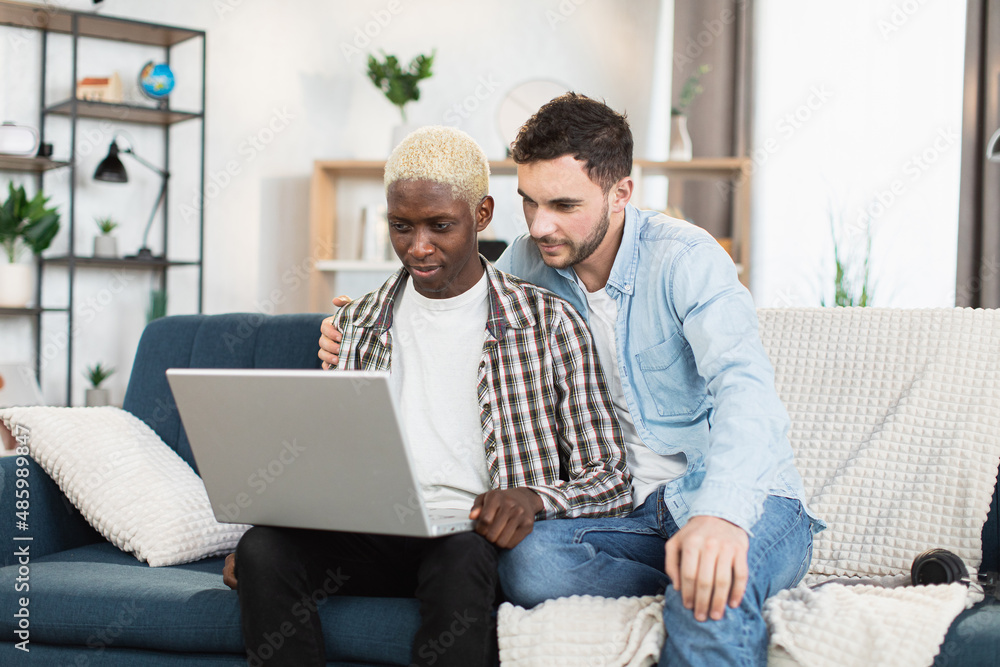 Multi ethnic couple of two gays using wireless laptop for video chat at home. Happy loving couple smiling and waving with hands while sitting together on couch. Multi ethnic couple of two gays using wireless laptop for video chat at home. Happy loving couple smiling and waving with hands while sitting together on couch.