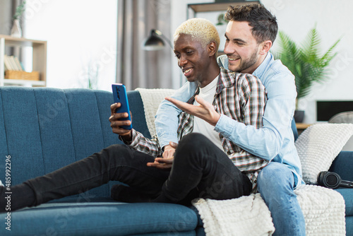 Beautiful and happy couple of multiracial gays sitting in embrace on comfy couch and using modern smartphone. Two loving people enjoying time spending together.
