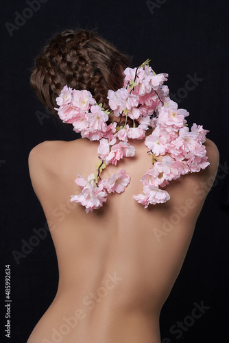 Fototapeta Naklejka Na Ścianę i Meble -  Back shot of a naked young girl with a branch of pink sakura. The girl is posing on the black backdrop.
