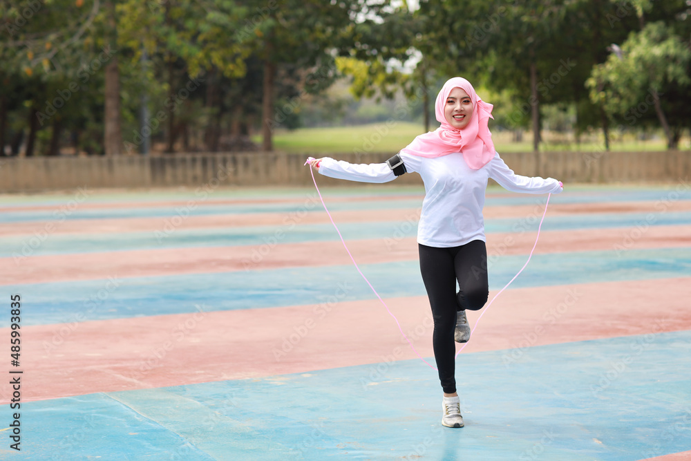 Athletic young asian woman in muslim sportswear standing and skipping rope outdoor for morning exercise. Active girl work out on biceps exercise with green tree background. Sport concept