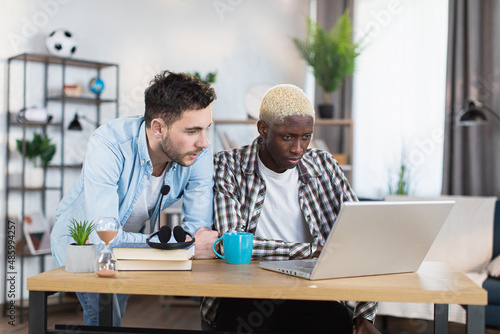 Multiracial same sex couple of two handsome gays using modern laptop ai living room. African american man sitting at desk while caucasian standing near.
