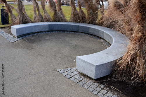 Japanese tall dry grasses for the winter tied in sheaves. in the arch of the flowerbed surround the granite bench in the park. The bench is bent like a horseshoe, with a smooth surface by the sidewalk photo