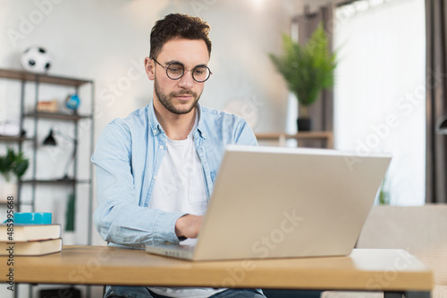 Attractive young man in eyewear and casual clothes sitting at domestic workplace and using modern laptop for work. Concept of people, technology and freelance.