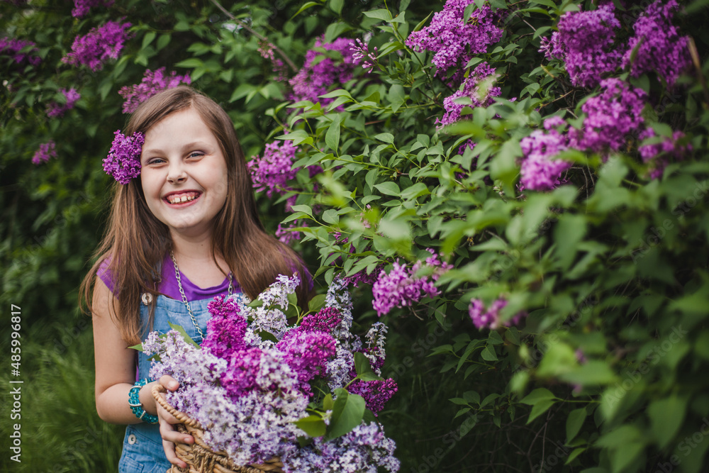 a teen laughing European girl in a denim jumpsuit holds a basket with lilacs in the garden near lilac bushes a teen laughing European girl in a denim jumpsuit holds a basket with lilacs in the garden near lilac bushes