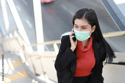 Young asian business woman in business black suit with protect mask for healthcare walking on footbridge public outdoor and using smartphone. New normal and social distancing concept