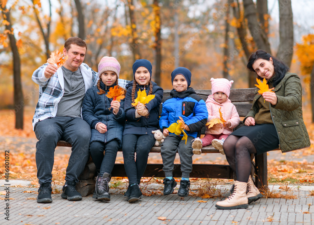 Fototapeta premium portrait of a family with children in an autumn city park - happy people sitting together on a wooden bench, posing against a background of beautiful yellow leaves