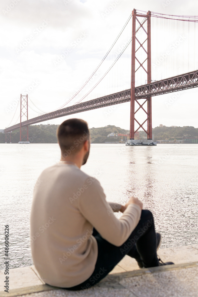 Unrecognizable man on his back sitting on the ground while looking towards the 25 april bridge in lisbon