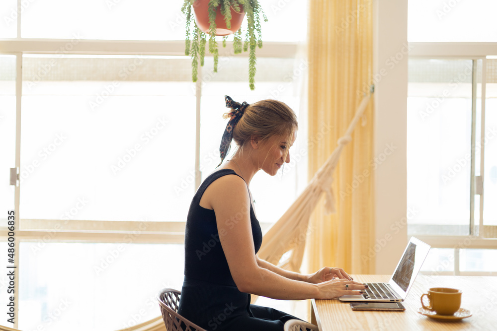 Young woman working at home in her kitchen with laptop and papers on kitchen wooden desk. Home office concept. Young woman working at home in her kitchen with laptop and papers on kitchen wooden desk. Home office concept.