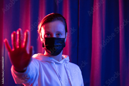 Young beautiful woman in a black medical mask and a white hooded sweater on a blue-violet background shows hand gestures during a coronavirus pandemic. Selective focus. Portrait