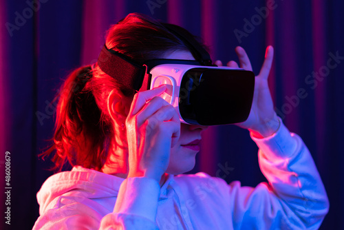 Young beautiful woman in virtual reality glasses and a white hooded sweater on a blue-violet background. Modern technologies. Selective focus. Portrait