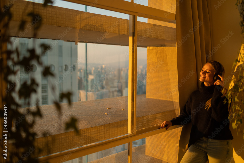 Side view of young woman listening to music with headphones in her ears by the window with a city view landscape in the background. High quality photo