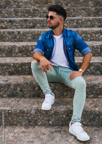 Full length shot of a cool style man with short hair wearing sunglasses sitting on a stairs and looking to the right. © anuatmoralesfoto