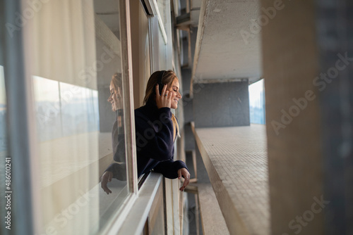 Side view of young woman listening to music with headphones in her ears by the window with a city view landscape in the background. High quality photo