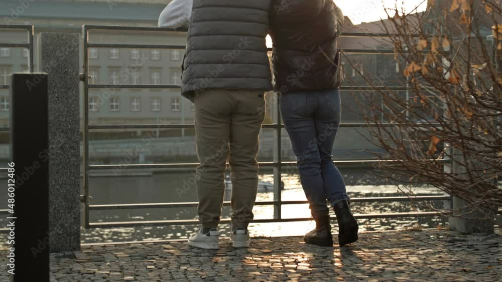 Man and a woman stand at the metal railing on the embankment and admire the view of the river and the city. Back view. Man and a woman stand at the metal railing on the embankment and admire the view of the river and the city. Back view.