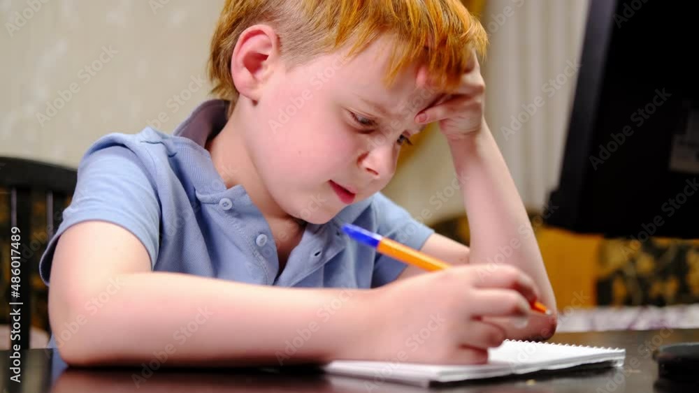 Primary school student diligently, tensely thinking while sitting at the table at home. Home schooling. Quarantine. A young red-haired boy. Primary school student diligently, tensely thinking while sitting at the table at home. Home schooling. Quarantine. A young red-haired boy.