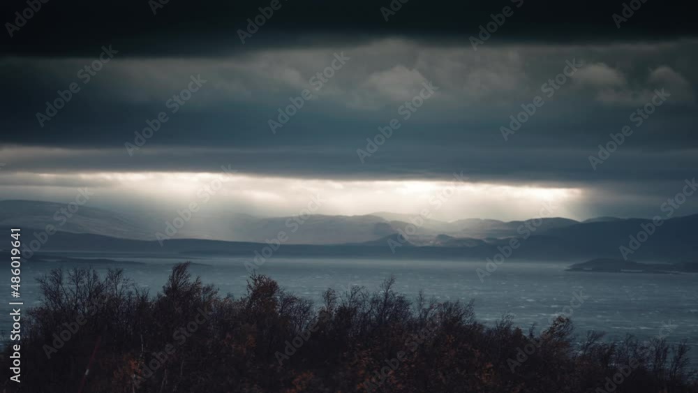 Sun rays pierce the thick menacing cloud cover above the fjord lighting the waters below. Windswept northern landscape in the foreground. Timelapse Sun rays pierce the thick menacing cloud cover above the fjord lighting the waters below. Windswept northern landscape in the foreground. Timelapse