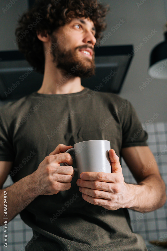 Close-up low-angle view of pensive young man holding in hand cup with morning coffee, thoughtful looking away, standing in kitchen with modern light interior, enjoying fresh warm drink at home office. Close-up low-angle view of pensive young man holding in hand cup with morning coffee, thoughtful looking away, standing in kitchen with modern light interior, enjoying fresh warm drink at home office.