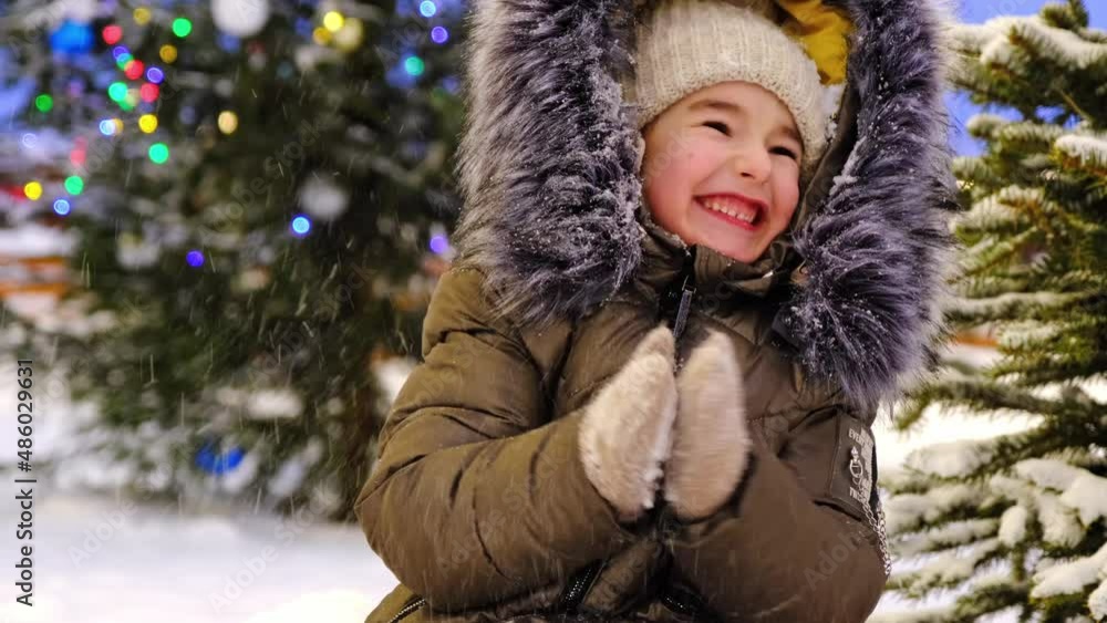 Portrait of happy girl in winter in fur hood against background of fairy lights, Christmas trees in snow in evening in winter outdoor. Play throws snow, claps her hands, laughs. Christmas, New Year Portrait of happy girl in winter in fur hood against background of fairy lights, Christmas trees in snow in evening in winter outdoor. Play throws snow, claps her hands, laughs. Christmas, New Year