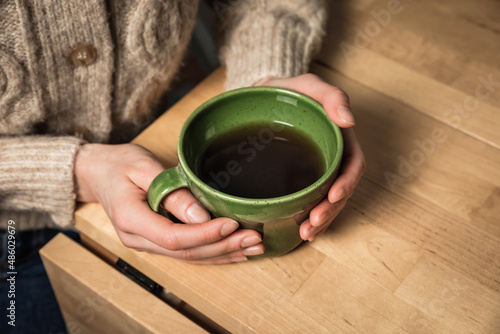 A caucasian woman in a cozy beige cardigan holding a green cup of tea in her hands, sitting at the table, a close-up image