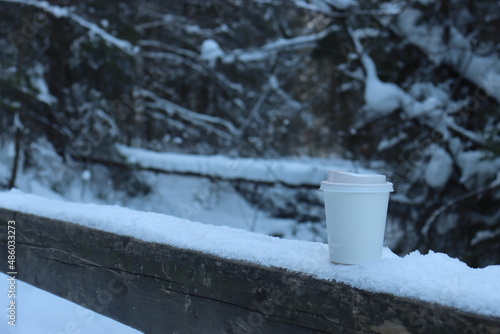 eco-friendly paper cup with a lid among the snow-covered dunes in the Alps. Stands on the railing of a wooden bridge passing through a mountain river February 2022