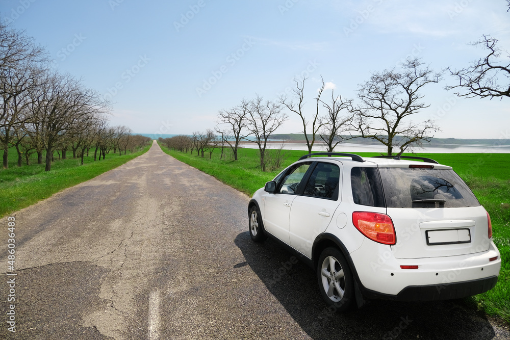 A white car after the rain stands on the side of a beautiful road that goes into the distance to the sea along a field and trees, auto travel