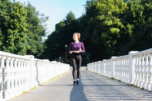 Sports, training, outdoor fitness. Slim smiling young woman in tracksuit running across bridge in park on sunny day and looking away. Smiling athletic caucasian sportswoman exercising outdoors © Sergio