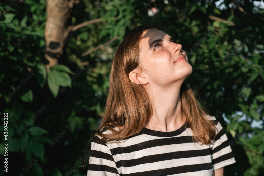 Portrait of young lovely woman with blue eyes and freckles in striped T-shirt and jeans against background of green foliage. Portrait of young lovely woman with blue eyes and freckles in striped T-shirt and jeans against background of green foliage.