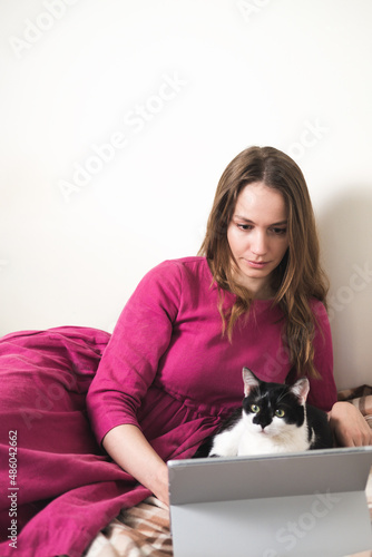 Young millennial woman in pink dress with long hair using a tablet and watcing movies with her black and white tuxedo cat at home.