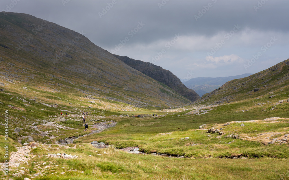 STYHEAD GILL, CUMBRIA, ENGLAND, UK - JULY 14, 2018: Distant Hikers walking along Styhead Gill below the summit of Base Brown. STYHEAD GILL, CUMBRIA, ENGLAND, UK - JULY 14, 2018: Distant Hikers walking along Styhead Gill below the summit of Base Brown.
