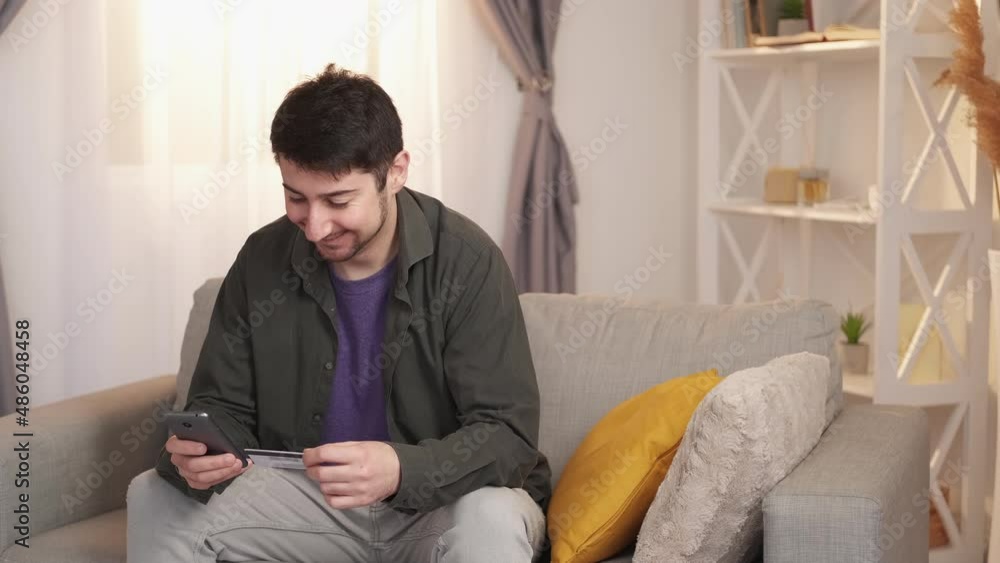 Online shopping. Internet payment. Phone banking. Joyful surprised man buying purchase on smartphone with credit card on sofa at home living room free space. Online shopping. Internet payment. Phone banking. Joyful surprised man buying purchase on smartphone with credit card on sofa at home living room free space.