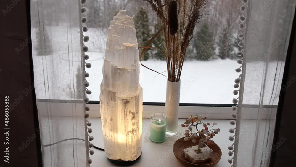 Switch on rough big selenite crystal tower pole lamp illuminated on home window sill, spiritual home decor accent. Winter forest on background.