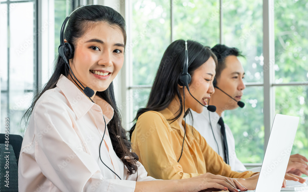 Focus Asian beautiful attractive female call center cheerful smiling with headsets, sitting, looking camera, monitoring with colleagues in office or agency at computer, giving service to customers. Focus Asian beautiful attractive female call center cheerful smiling with headsets, sitting, looking camera, monitoring with colleagues in office or agency at computer, giving service to customers.