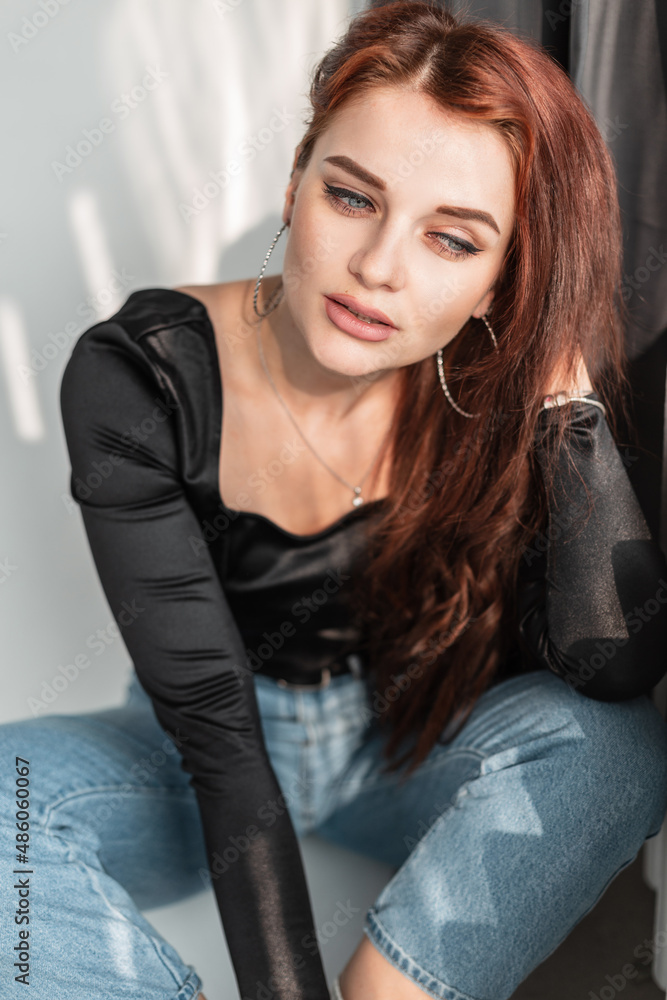 Beautiful young girl in a fashionable denim outfit with a black T-shirt and blue jeans is sitting near the window with sunlight indoors Beautiful young girl in a fashionable denim outfit with a black T-shirt and blue jeans is sitting near the window with sunlight indoors