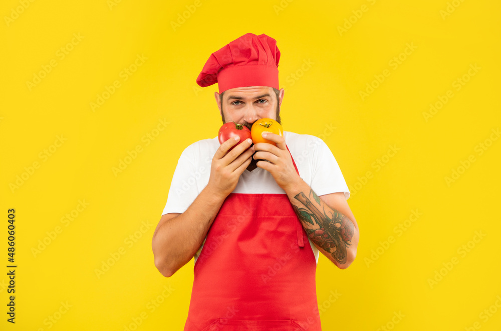 Serious guy in cooking apron and toque smelling tomatoes yellow background, chef Serious guy in cooking apron and toque smelling tomatoes yellow background, chef