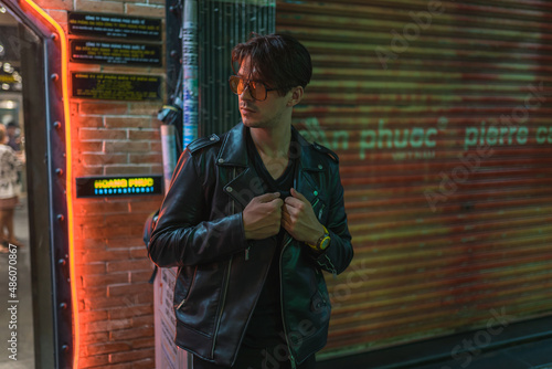 Portrait of young stylish brunet caucasian man in black leather jacket and yellow sunglasses. Standing on the street near the wall in evening. 