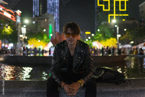 Portrait of young stylish brunet caucasian man in black leather jacket and yellow sunglasses. Sitting outdoors at night and looking at camera 