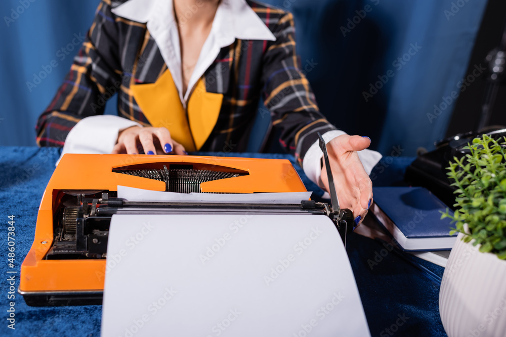 cropped view of stylish woman near vintage typewriter with blank paper on blue background