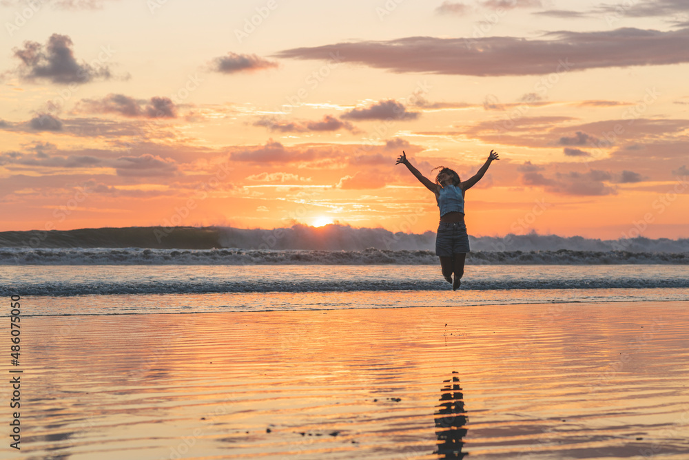 Latin woman enjoying the beach in a beautiful sunset, jumping with open arms in happiness. 