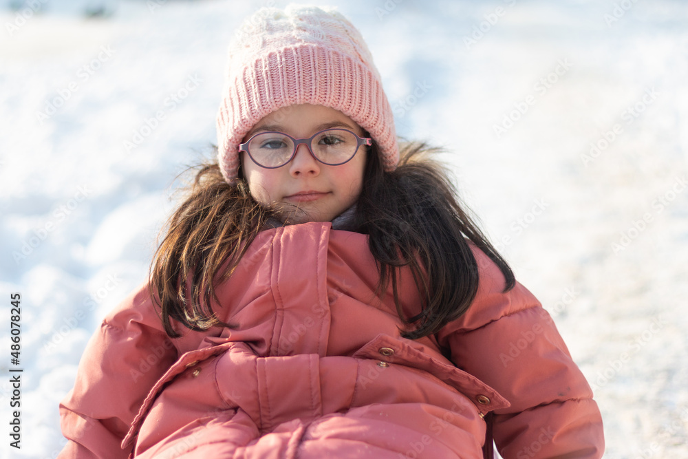 A small child in winter clothes and a knitted hat and glasses looks ahead while sitting on a sled. Winter photo session outdoors A small child in winter clothes and a knitted hat and glasses looks ahead while sitting on a sled. Winter photo session outdoors