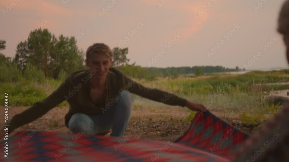 Part of romantic love story, young couple set up picnic blanket in forest, next to beach. Loving and romantic setting for a date. Authentic millennial youth interact outdoors Part of romantic love story, young couple set up picnic blanket in forest, next to beach. Loving and romantic setting for a date. Authentic millennial youth interact outdoors
