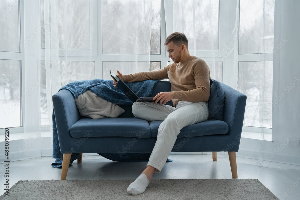 A young man works at home on the couch with his laptop. A man with a mobile phone and a laptop is sitting on a blue sofa against the background of a window.