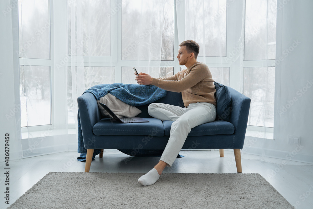 A man with a mobile phone and a laptop is sitting on a blue sofa against the background of a window. A young man works at home on the couch with his laptop