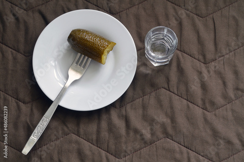 Pickled cucumber on a fork on a white saucer, a glass of vodka on a brown background.