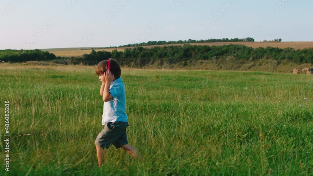In a large green field cute little boy take his red headphones and listen music chilling walking through the grass in front of the camera. Shot on ARRI Alexa Mini. In a large green field cute little boy take his red headphones and listen music chilling walking through the grass in front of the camera. Shot on ARRI Alexa Mini.