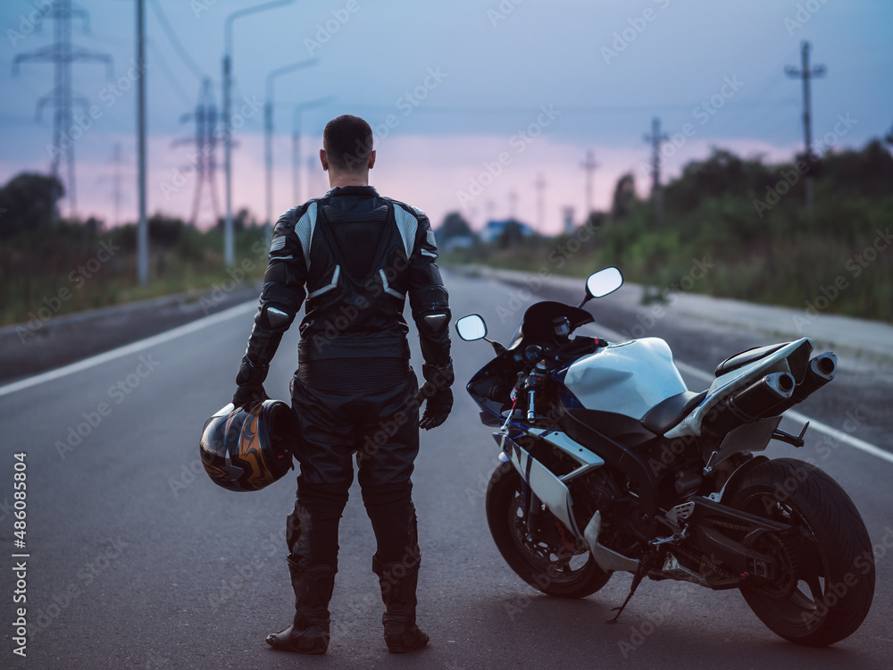 Fototapeta premium A biker stands next to a motorcycle on an asphalt road and looks into the distance. Summer sunset