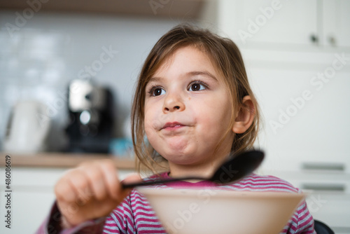Close up of preschool girl portrait with funny expression eating soup at home 