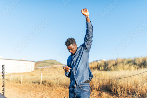 young african american man raising fist in victory sign looking at smartphone on sunny day photo