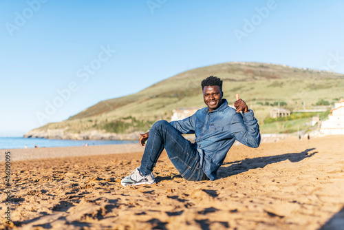 young african american man sitting on the beach smiling raising thumb up on a sunny day. approval concept © Patricia