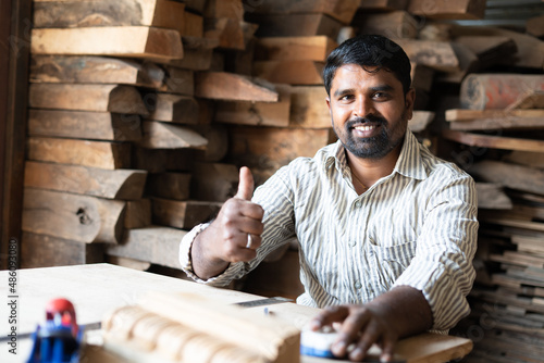 Fotografija Smiling carpenter showing thumbs up by looking at camera while working at shop - concept of approve, promotion, artisan and wood working
