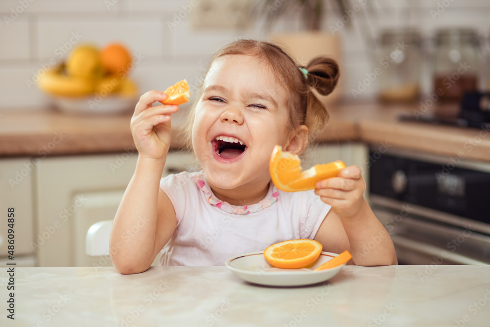 A happy girl 2-3 years old in the kitchen at home or in kindergarten eats delicious fruits, sweet oranges.